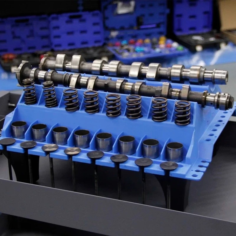 Variklio techninės priežiūros darbo stalas: plastikinė plokštė įsiurbimo/ išmetimo dalių išdėstymui — cilindro galvos varžtai, vožtuvo svirčių rankos, viršutinės talpyklos ir vožtuvo spyruoklių vietos